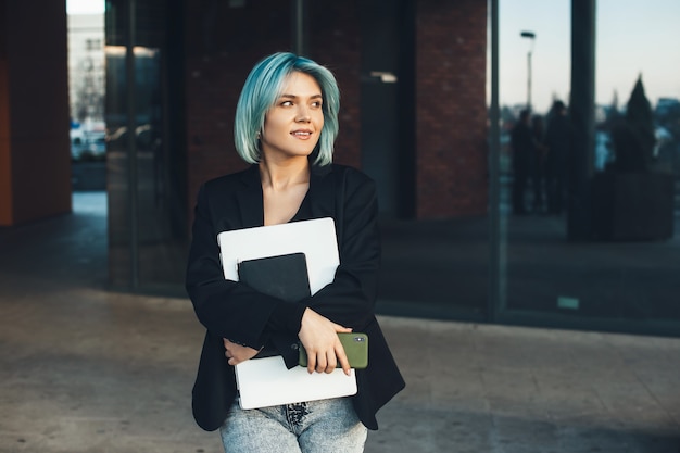 Charming caucasian businesswoman with blue hair is looking away while holding a laptop and phone in front of a building