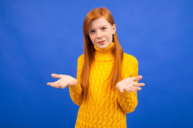 Charming caucasian red-haired girl in a yellow sweater waving her arms on a blue studio background.