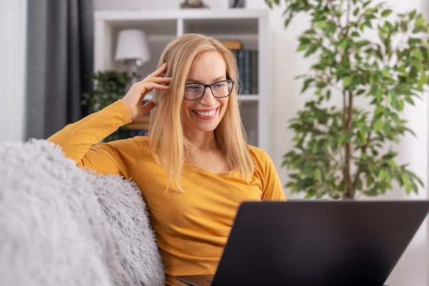 Charming caucasian woman in casual outfit and eyeglasses sitting on couch and using wireless laptop Technology people and domestic life concept