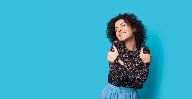 Charming curly haired woman is embracing herself on a blue wall smiling near free space