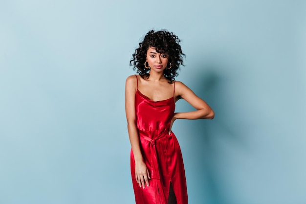 Charming curly young woman in red dress looking at camera Studio shot of confident mixed race girl isolated on blue background