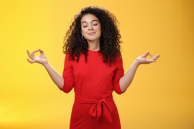 Charming dreamy and happy peaceful woman in red dress with curly hair standing in lotus pose with mu...