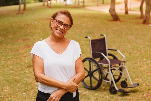 휠체어와 함께 서있는 공원에서 웃는 매력적인 노인 여성은