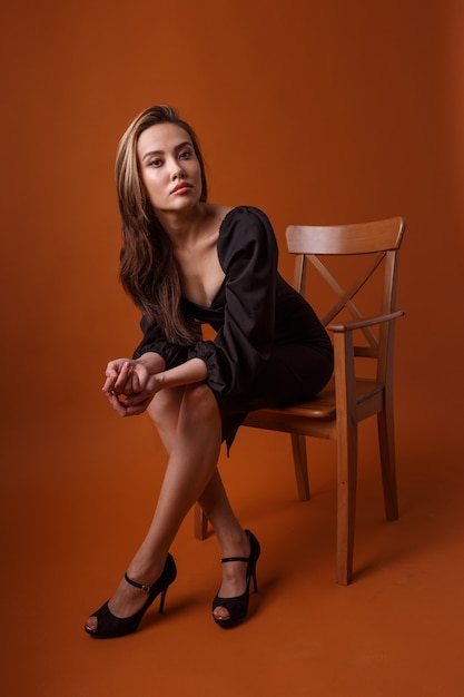 Charming elegant fashion model wearing black dress, high heel shoes posing on orange background