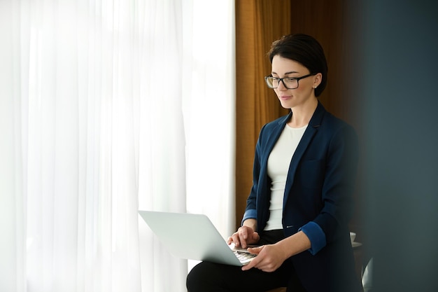 Charming elegant young prosperous pensive entrepreneur, business woman typing on a laptop a progress report, sitting in the edge of a desk in a hotel room during a business trip