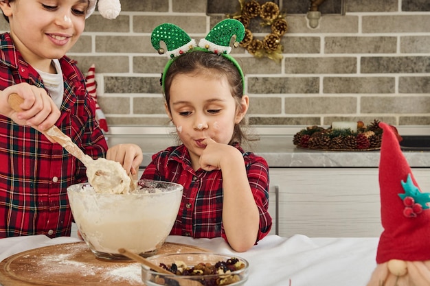 매력적인 유럽 어린이들이 집에서 함께 크리스마스 쿠키를 요리하고 있습니다. 귀여운 여동생이 반죽에 손을 올려 맛보면 사랑스러운 소년이 나무 숟가락으로 반죽을 반죽한다