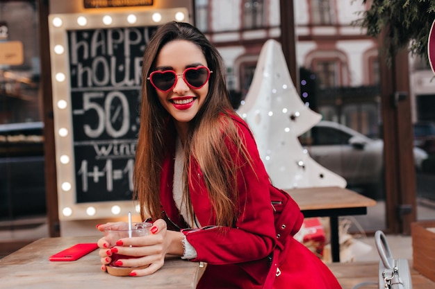 Charming european girl in trendy red attire spending time in cafe. Romantic white lady with straight hairstyle relaxing and drinking tea.