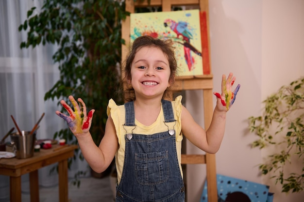 A charming European preschool age child cute 4 years old girl smiles sweetly standing at wooden easel and showing her hands stained on colorful in paints for creativity to the camera