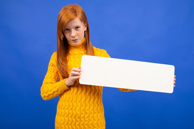 Charming european red-haired teenage girl holding a banner from a sheet of paper for advertising on blue