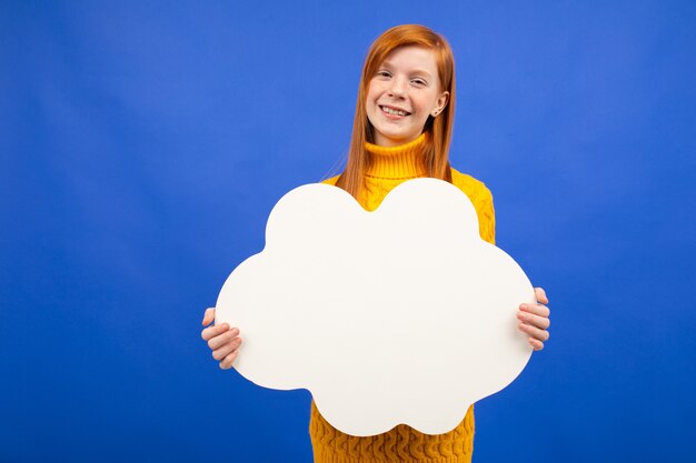 Charming european red-haired teenage girl holding a banner from a sheet of paper with a mockup for advertising on a blue studio background