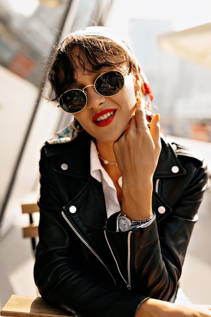 Charming excellent lady in sunglasses with red lips wearing black jacket and bright scarf on the head is sitting outdoor on summer terrace in sunshine