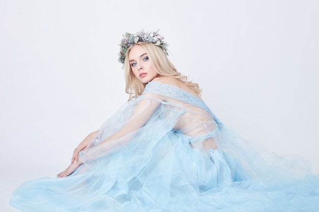 Charming fairy woman in blue ethereal dress and wreath on head on white background, gentle mysterious blonde girl with perfect skin and makeup. Cleanliness, body care and skin