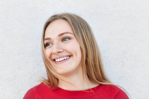 Charming female model with straight fair hair looking with her blue eyes aside having broad smile demonstrating her white teeth. Good-looking woman in red sweater with dimples on cheeks smiling