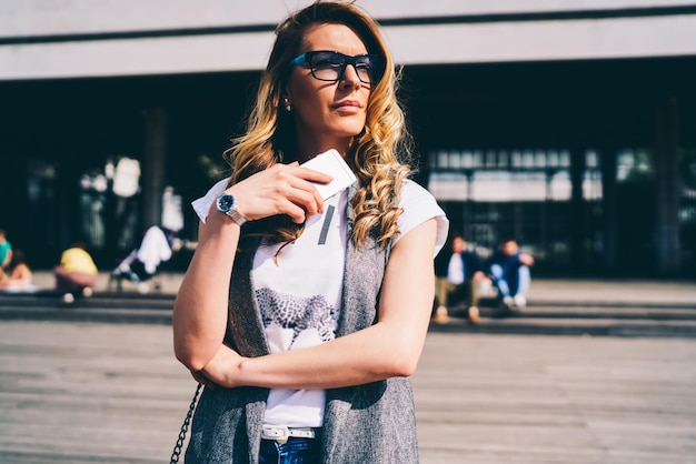 Charming female person in spectacles enjoying leisure time while strolling at sunny urban setting