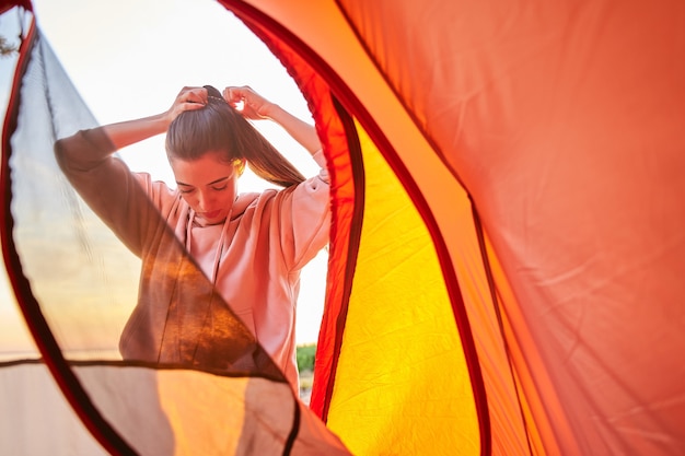 Charming female traveler in hoodie putting her hair up in ponytail while standing neat touristic tent