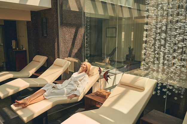 Photo charming female wearing white bathrobe while lying on the sun bed in spa resort hotel