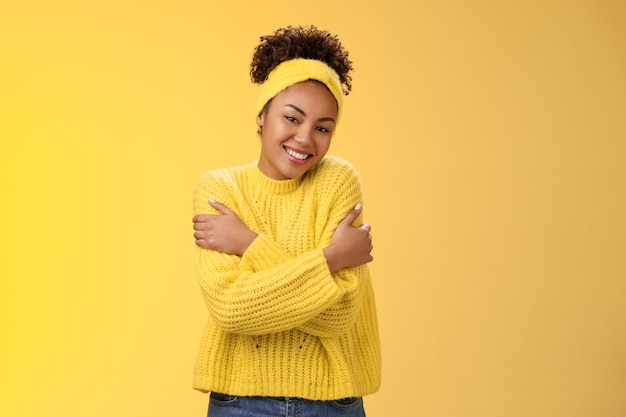 Charming feminine african-american woman lovely tender smile tilting head hugging herself feel comfort love gently cuddling shoulders wearking comfy sweater, grinning happily yellow background.