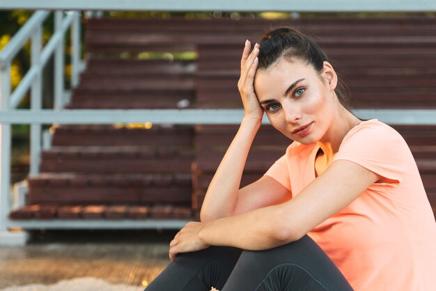 charming fitness girl in sportswear looking at front and propping up her head while sitting at sports ground