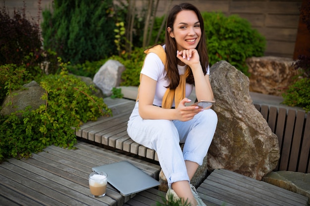 カジュアルな表情で魅力的な無料の若い女性は、夏の公園に座ってコーヒーを飲んでいます。