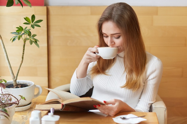 Charming fun-loving girl takes sips of tea, reads action packed book, focuses all attention on plot. Young model sits near green plants and hot coffee on table.