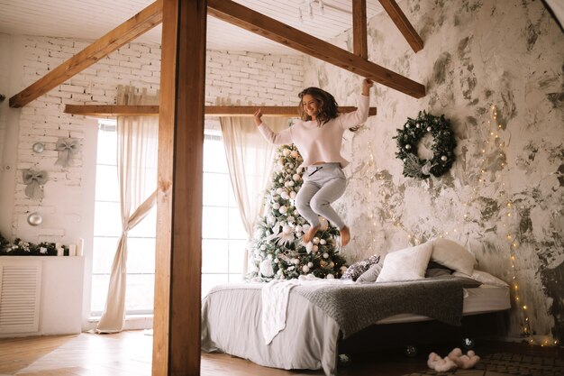 Charming girl dressed in white sweater and pants jumps on the bed with gray blanket and white pillows in a cozy decorated room with a New Year tree . .