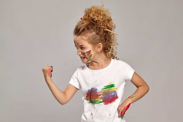 Charming girl having a brush in her lovely haircut, wearing in a white smeared t-shirt. She is posing with a painted hands and cheeks, showing a fist to someone and looking angry, on a gray background