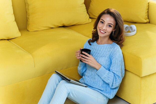 Charming girl holds her tablet computer in hands, sitting on the floor near modern yellow couch and drinks coffee in the modern stylish kitchen. Young cheerful woman having rest at cozy home.