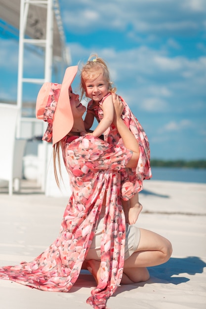 明るい夏のサンドレスを着た魅力的な女の子が、小さな娘と一緒に砂浜を歩きます。暖かく晴れた夏の日を楽しんでいます。