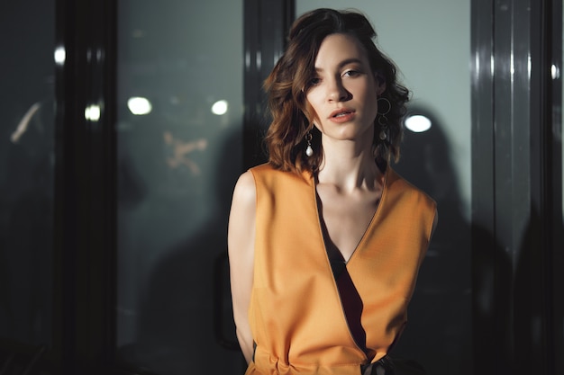 Charming girl posing in front of glass doors with reflection of lights on it