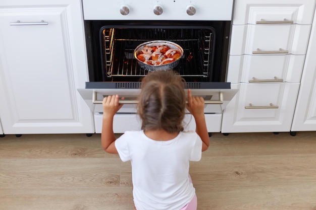 Charming girl sitting near gas stove on floor in kitchen and opening oven curious female child waiting for cake or pie baking in stove homemade pastry