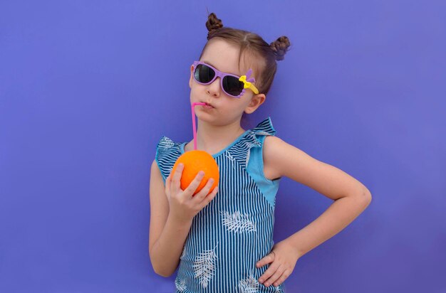 Charming girl in summer glasses drinks orange juice on a purple background Healthy lifestyle Copy spaces