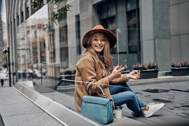 Charming girl. Through the glass case view of attractive young woman in hat and coat looking at camera and smiling while using her smart phone