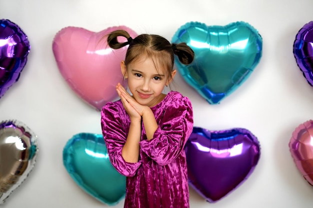 Charming girl with ponytails in a velvet purple dress stands against the background of multicolored balloons in the shape of a heart for valentine's day, cute pressing her hands to her cheeks