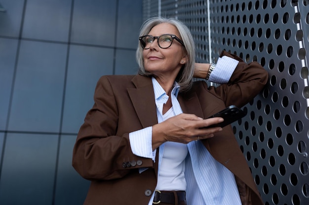 Foto affascinante donna di mezza età dai capelli grigi e ben curata con gli occhiali vestita con una giacca marrone per strada