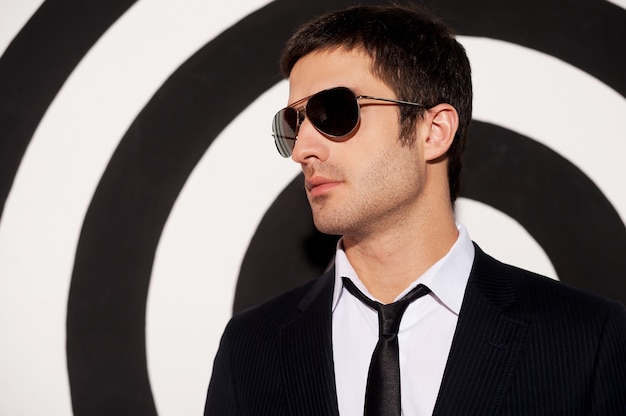 Charming handsome. Portrait of handsome young man in formalwear and sunglasses looking away while standing against black and white background