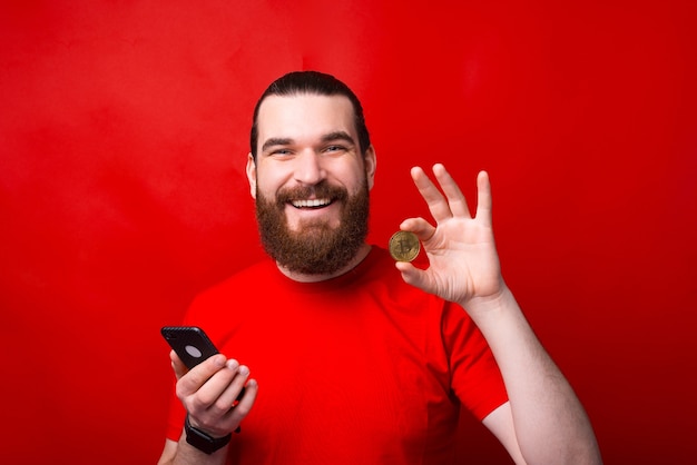 Charming handsome young bearded man holding smartphone and showing bitcoin over red wall