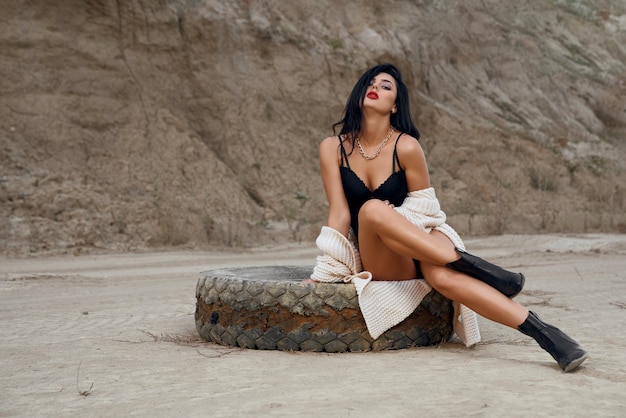 Charming hot woman dressed in trendy black lingerie and knitted cardigan sitting on old tyre among sand quarry