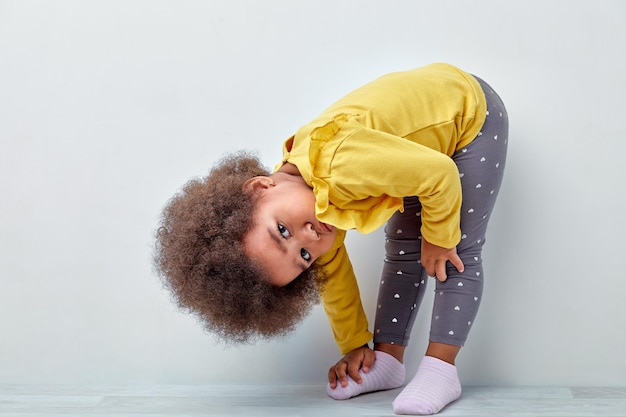 Photo charming kid girl of african appearance stand with head bowed looking at camera, cute child in casual wear posing at camera, lovely mixed race child girl with fluffy wavy hair