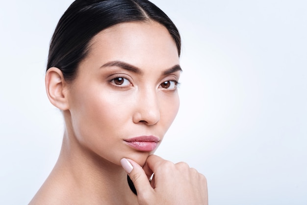 Charming lady. The close up of a pretty dark-haired young woman touching her chin and  while posing against a white wall