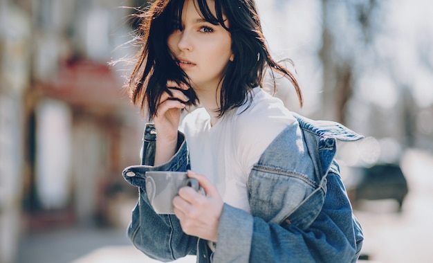 Charming  lady posing outside being dressed in a blue jacket and holding a cup of tea