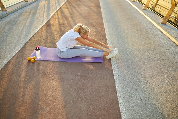 Charming lady in sportswear doing stretching exercise while sitting on yoga mat outdoors