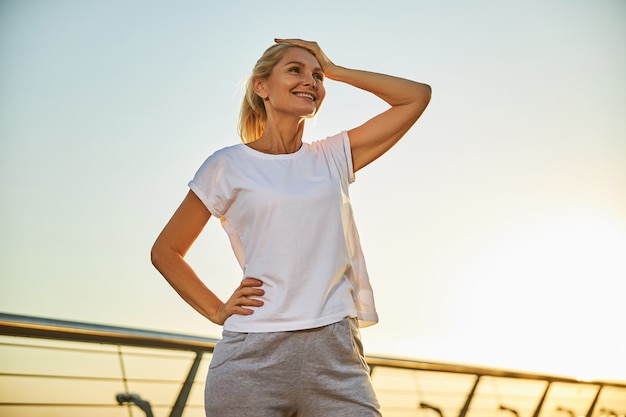 Charming lady in white shirt placing hand on her head and smiling while spending time outdoors