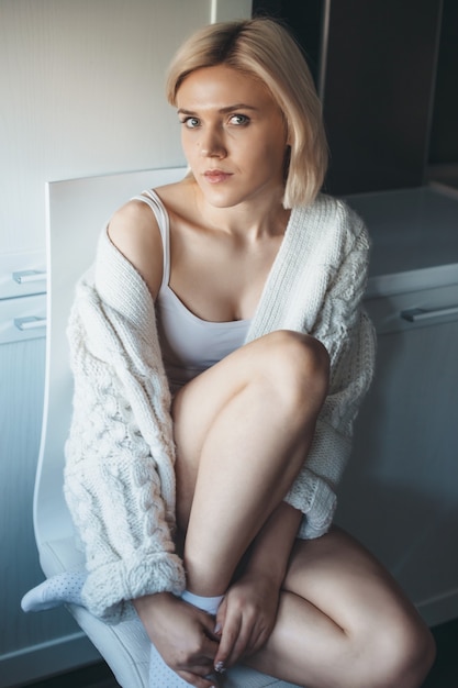 Charming lady with blonde hair and knitted sweater is sitting on the chair in the kitchen