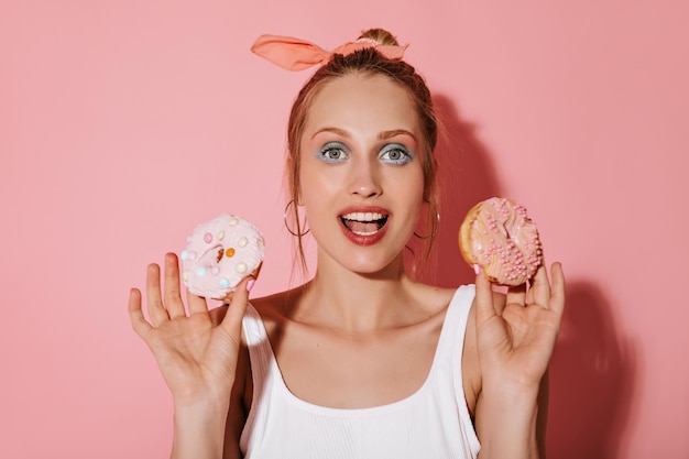 Charming lady with blonde hair and trendy makeup in white swimsuit looking into camera and holding donuts on isolated background