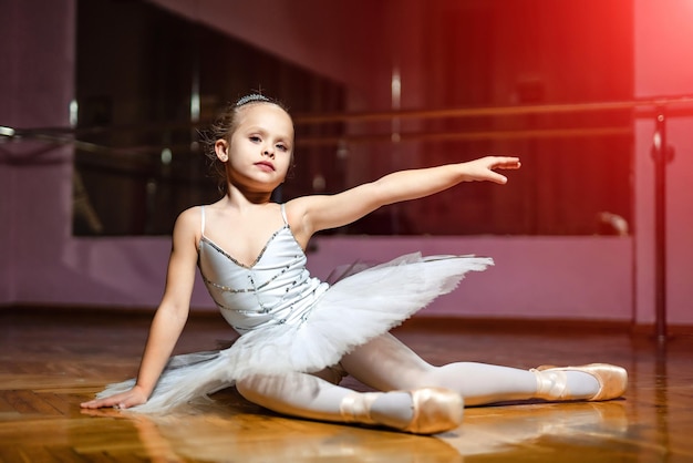 Charming little ballet dancer in white tutu sitting on the floor and poses to camera on the studio background Sweet ballerina sitting on wooden floor in a dance studio