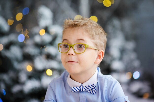 Charming little boy is sitting at home, snowy winter decorated tree on background