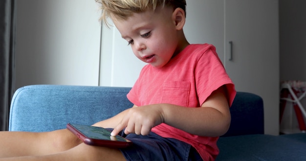 Очаровательный маленький мальчик сидит на синем диване у себя дома и играет в игру на смартфоне