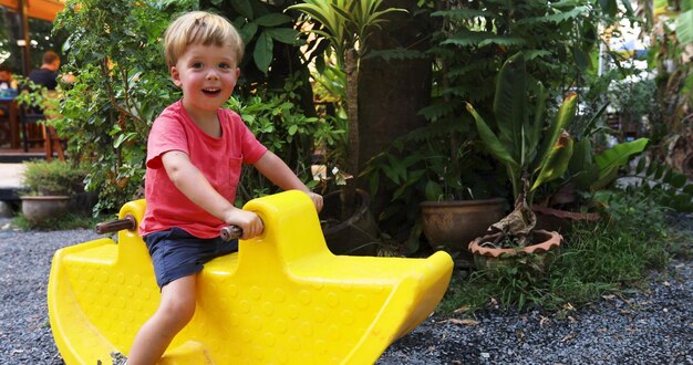 Charming little boy sitting on yellow toy ride and having fun on resort playground smiling at camera 