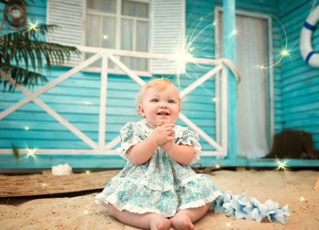 Charming little fair-haired half-year-old sorceress girl in a blue dress sits against the background of a country house. The concept of adorable and naive babies