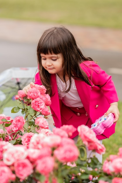 밝은 분홍색 양복을 입은 매력적인 어린 소녀가 장미 덤불 카드 배너를 킁킁